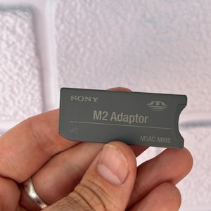 Op de afbeelding: Een grijze Sony M2 Adapter met de tekst "M2 Adaptor" en "MSAC-MMS" erop gedrukt. De adapter is rechthoekig met een gebogen rand en een kleine pijl. Het Sony-logo is bovenaan zichtbaar.