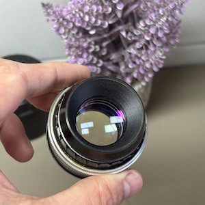 Può includere: Un obiettivo per fotocamera vintage con un corpo in metallo nero e argento. L'obiettivo ha un elemento in vetro trasparente con riflessi. L'obiettivo è tenuto in mano, con uno sfondo sfocato di fiori viola.