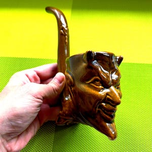 May include: A brown ceramic pipe in the shape of a devil's head, featuring horns and a smiling expression. The pipe has a curved stem and is held in a hand.