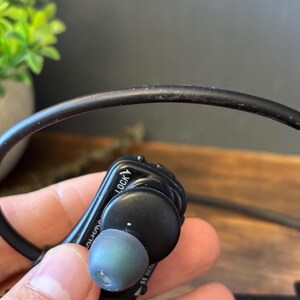 May include: A black in-ear headphone with a flexible black cable and a grey silicone ear tip. The device features a prominent black housing with the word LOCK printed in white lettering near the top edge and a curved design for ear placement.