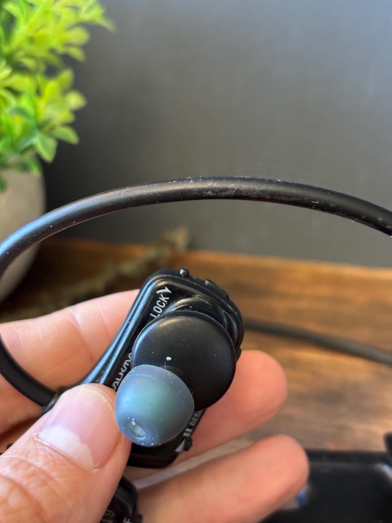 May include: A black in-ear headphone with a flexible black cable and a grey silicone ear tip. The device features a prominent black housing with the word LOCK printed in white lettering near the top edge and a curved design for ear placement.