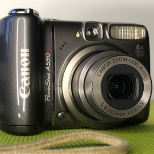 以下が含まれることがあります： 濃いグレーのCanon PowerShot A590 ISデジタルカメラ。カメラには「Canon Zoom Lens 4x IS」と測定の詳細が記載されたシルバーのレンズリングが付いています。また、フラッシュとリストストラップも付いています。