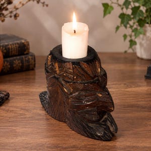 May include: A dark brown, hand-carved wooden candle holder shaped like a human face with a prominent nose and textured features. The holder has a circular opening at the top containing a single, lit, cylindrical white wax candle with a visible flame.