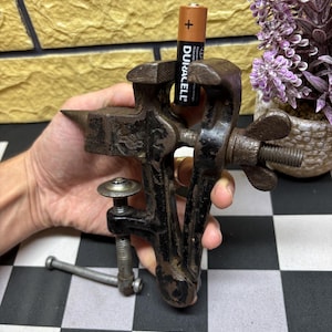 Puede incluir: Un pequeño tornillo de banco de metal negro vintage con un mecanismo de tornillo y una abrazadera. Una pila Duracell AA se sostiene en el tornillo de banco. El tornillo de banco está sobre una superficie a cuadros en blanco y negro.