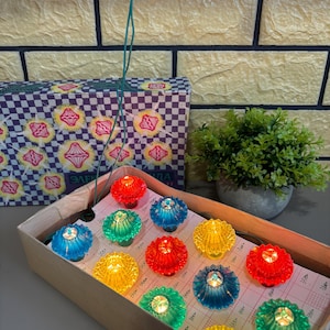 Może przedstawiać: Kolekcja kolorowych, zabytkowych lampek choinkowych w kartonowym pudełku. Lampki mają kształt kwiatów i są w kolorze czerwonym, żółtym, zielonym i niebieskim. W tle pudełko z kwiatowym wzorem.