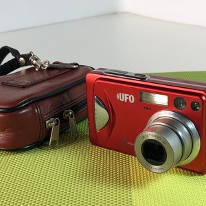 以下が含まれることがあります： 銀色のレンズと黒い革製のキャリングケースが付いた赤いデジタルカメラ。カメラの前面には「UFO」という文字が印刷されています。