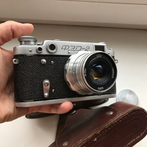 Może przedstawiać: Srebrno-czarny aparat fotograficzny 35 mm z obiektywem oznaczonym "ФЭД-2" i "Jupiter-8 50mm 1:2". Aparat jest trzymany w dłoni i leży na brązowym sk&oacute;rzanym etui.