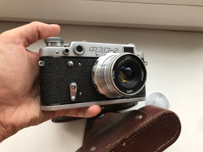 Może przedstawiać: Srebrno-czarny aparat fotograficzny 35 mm z obiektywem oznaczonym "ФЭД-2" i "Jupiter-8 50mm 1:2". Aparat jest trzymany w dłoni i leży na brązowym sk&oacute;rzanym etui.