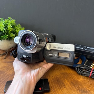 May include: A grey and black Sony Handycam Vision video camera with a flip-out screen. The device features a Sony Video Lens with 20x optical zoom, SteadyShot technology, and is labeled as a CCD-TRV408E PAL model that records in video Hi8 format.