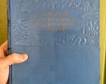 Puede incluir: Un libro de tapa dura azul con ilustraciones en relieve de varios alimentos a lo largo del borde. La portada presenta el texto cirílico "КНИГА О ВКУСНОЙ И ЗДОРОВОЙ ПИЩЕ" en letras doradas centradas dentro del marco decorativo.