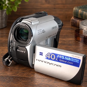 May include: A silver and black Sony Handycam DCR-DVD109 camcorder with a Carl Zeiss Vario-Tessar lens. The flip-out screen displays 40x Optical Zoom and 5.1ch Surround Recording text. The device features a black wrist strap and silver body accents.