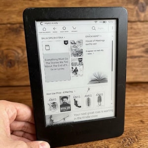 Könnte beinhalten: Ein schwarzer E-Reader mit einem rechteckigen Bildschirm, der eine digitale Bibliotheks-Benutzeroberfläche anzeigt. Der Bildschirm zeigt Buchcover, Titel wie Everything Must Go und Gulag sowie Navigationssymbole. Das Gerät hat einen schwarzen Rahmen.