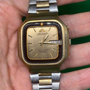 May include: Vintage Orient Crystal 21 Jewels watch with a gold-tone case and a two-tone silver and gold band. The watch face is gold with black accents, featuring the day and date. The watch is a classic timepiece.
