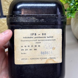 Op de afbeelding: Een zwarte, rechthoekige container met afgeronde randen. Op de voorkant staat een label met de tekst "IPB - 80" en tekst in meerdere talen. Het label bevat ook verschillende regels met nummers en de tekst "LÉČIVA - NARODNI PODNIK - PRAHA."