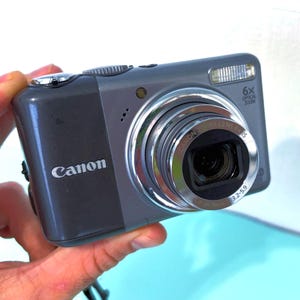 May include: A grey Canon digital camera with a silver lens and a built-in flash. The camera has a 6x optical zoom and the Canon logo is visible on the front. The camera is held in a hand.