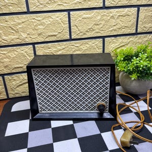 May include: A vintage black radio with a white speaker grill and a tuning knob. The radio has a retro design and is placed on a black and white checkered surface. A brown electrical cord and plug are visible.