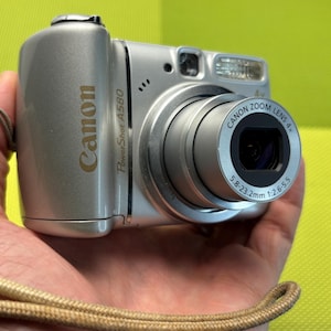 以下が含まれることがあります： 4倍ズームレンズを搭載したシルバーのキヤノンPowerShot A580デジタルカメラ。レンズ鏡筒にはCanon Zoom Lens 4x 5.8-23.2mm 1:2.6-5.5と印字されています。カメラ本体にはキヤノンのロゴとPowerShot A580の文字があり、ベージュのリストストラップが付いています。