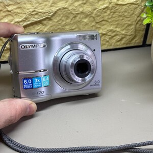 以下が含まれることがあります： 6.0メガピクセルの解像度と3倍光学ズームを備えた、シルバーのオリンパスE-170デジタルカメラ。「OLYMPUS LENS」の文字が入ったレンズと内蔵フラッシュがあります。カメラは手で持たれており、灰色のストラップが付いています。