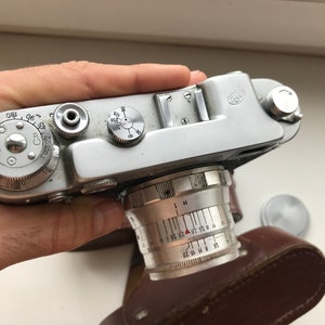 Może przedstawiać: Srebrny, zabytkowy aparat fotograficzny w sk&oacute;rzanym etui. Aparat ma obiektyw z skalą głębi ostrości i srebrny pokrętło z liczbami i symbolami. Aparat ma srebrny korpus i czarny sk&oacute;rzany pasek.