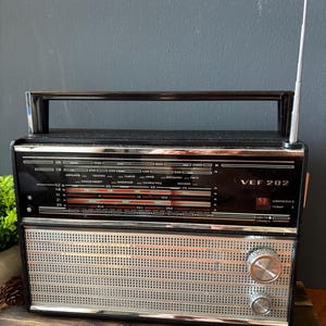 Könnte beinhalten: Ein schwarzes VEF 202 Kofferradio mit silbernem Metall-Lautsprechergitter, einem Tragegriff an der Oberseite und einer langen Teleskopantenne. Die Frontplatte zeigt eine Frequenzskala mit kyrillischer Schrift sowie zwei runde Drehregler.