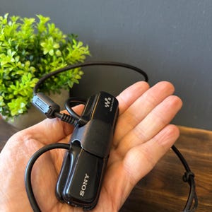 May include: A black Sony Walkman digital music player featuring a compact, rectangular design with the Sony logo and Walkman branding printed on the front. The device includes an attached black USB cable with a ferrite core and a standard USB connector.
