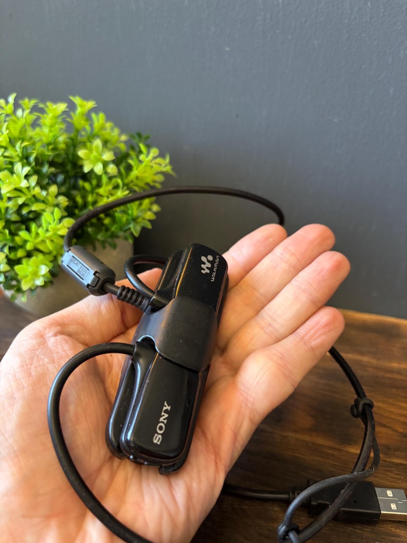 May include: A black Sony Walkman digital music player featuring a compact, rectangular design with the Sony logo and Walkman branding printed on the front. The device includes an attached black USB cable with a ferrite core and a standard USB connector.