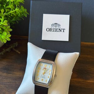 May include: An Orient wristwatch with a rectangular silver-tone case, a white dial featuring gold-colored Arabic numerals, and a black leather strap. The watch is displayed on a white cushion in front of a black box with the Orient logo printed on it.