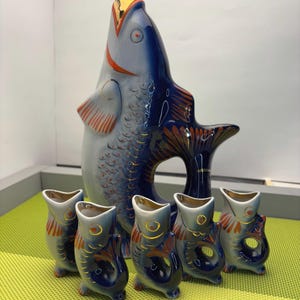 Puede incluir: Una botella de licor de porcelana azul y blanca con forma de pez con una tapa dorada y seis vasos de chupito a juego. La botella y los vasos están decorados con detalles rojos y dorados.