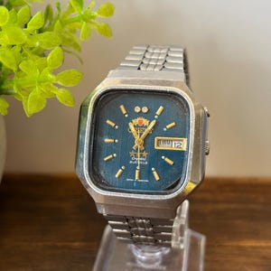 May include: Vintage Orient watch with a silver-tone metal band and a square face. The watch face is a deep teal color with gold-tone hands and markers. The day and date are displayed in a small window. The watch is displayed on a clear stand.