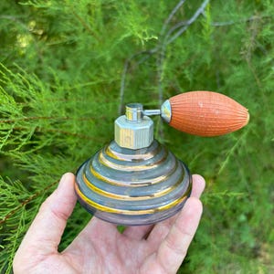 May include: Vintage perfume bottle with a silver atomizer and a bulb-shaped orange sprayer. The glass bottle is dark with gold and silver stripes. The bottle is held in front of a green leafy background.