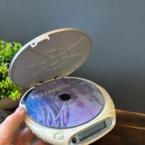 May include: A portable white CD player with an open lid, featuring a circular digital display and control buttons. Inside the player sits a compact disc with the text KLUBBHEADS printed on the surface. The device has a rounded design.