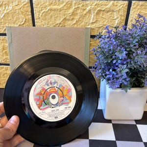 以下が含まれることがあります： クイーンのカラフルなラベルが付いた黒いビニールレコード。レコードは手で持たれています。ラベルには「WHITE MAN」と「EMI 2565」の文字があります。紫色の花が入った小さな白い正方形のポットが背景にあります。