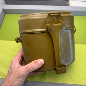 May include: A green metal camping mess kit with a folding handle. The kit has two nesting pots and a lid.