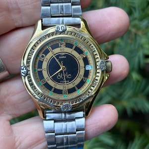 May include: A gold-toned wristwatch with a black face and silver strap. The watch face displays the word "Style" and numbers from 1 to 60. The watch has a gold bezel with black and green details and a silver strap.