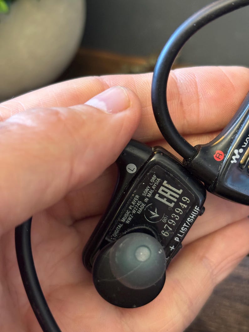 May include: A black Sony Walkman digital music player, model NWZ-W274S, featuring an in-ear design with a wrap-around band. The device displays the text 'DIGITAL MUSIC PLAYER', 'NWZ-W274S', 'SONY', 'MADE IN MALAYSIA', 'EAC', '6793949', and 'P.LIST/SHUF'.
