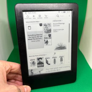Puede incluir: Un lector de libros electrónicos Kindle negro que muestra una biblioteca digital. La pantalla muestra portadas y títulos de libros, incluyendo "Everything Must Go" y "House of Meetings". El dispositivo se sostiene contra un fondo verde.