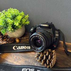 May include: A black Canon EOS Rebel XT digital camera with an attached Canon Zoom Lens EF 38-76mm 1:4.5-5.6. The camera features a black neck strap with the white text Canon and EOS DIGITAL printed on it, positioned near a pinecone.