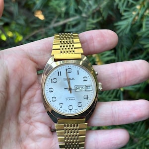 Op de afbeelding: Een goudkleurig SUVA automatisch polshorloge met een zilveren wijzerplaat, zwarte cijfers en wijzers. Het horloge toont de dag en datum, met een gouden mesh band. Het horloge wordt tegen een achtergrond van groen gebladerte gehouden.
