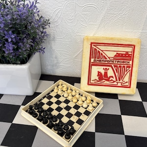 Op de afbeelding: Een vintage schaakspel met zwarte en witte stukken op een schaakbord. De set zit in een crèmekleurige koffer met rode tekst en afbeeldingen. Een kleine witte vierkante pot met paarse bloemen staat op de achtergrond.