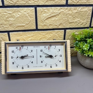 May include: A vintage chess clock with a cream-colored frame and two circular clock faces. The clock face has black numbers and hands, with red indicator needles. The clock is labeled "Made in USSR".