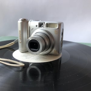 以下が含まれることがあります： 4倍光学ズームレンズ、800万画素、ベージュのリストストラップを備えたシルバーのキヤノンPowerShotデジタルカメラ。レンズ鏡筒にはCanon Zoom Lens 4x 5.8-23.2mm 1:2.6-5.5と刻印されています。カメラには内蔵フラッシュが搭載されています。