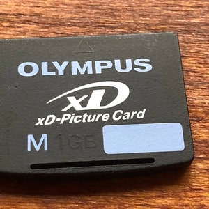 Puede incluir: Tarjeta xd-Picture Olympus negra con texto blanco y azul claro. La tarjeta tiene el texto "OLYMPUS", "xD", "xd-Picture Card", "M 1GB". La tarjeta es rectangular y está sobre una superficie de madera.