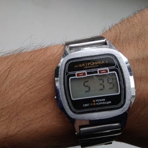 Puede incluir: Un reloj digital plateado con una esfera negra y una correa de metal. La esfera del reloj muestra la hora como 5:39. El reloj está etiquetado como "Электроника 5" y tiene botones para "свет" (luz), "режим" (modo) y "коррекция" (corrección).