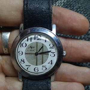 Puede incluir: Un reloj de pulsera plateado con una esfera blanca y una correa de cuero negra. El reloj tiene un patrón de cuadrícula en la esfera y agujas negras.