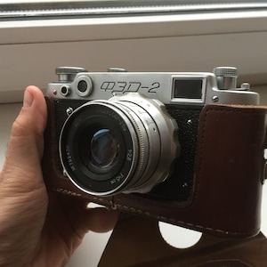 Puede incluir: Una cámara de película de 35 mm plateada y negra con una funda de cuero. La cámara tiene un objetivo con el texto "Индустар-28 2.8cm" impreso en él. La cámara está etiquetada como "ФЭД-2".