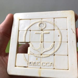 以下が含まれることがあります： 中央に錨と「ODESSA」の文字が描かれた円形のデザインが施された白いプラスチック製のジグソーパズルです。