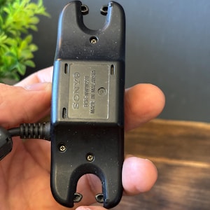 May include: A black Sony BCR-NWW270 charging cradle for a wearable device. The rectangular plastic unit features two U-shaped cutouts at the top and bottom, exposed screws, and text reading SONY BCR-NWW270 MADE IN MALAYSIA on the center panel.