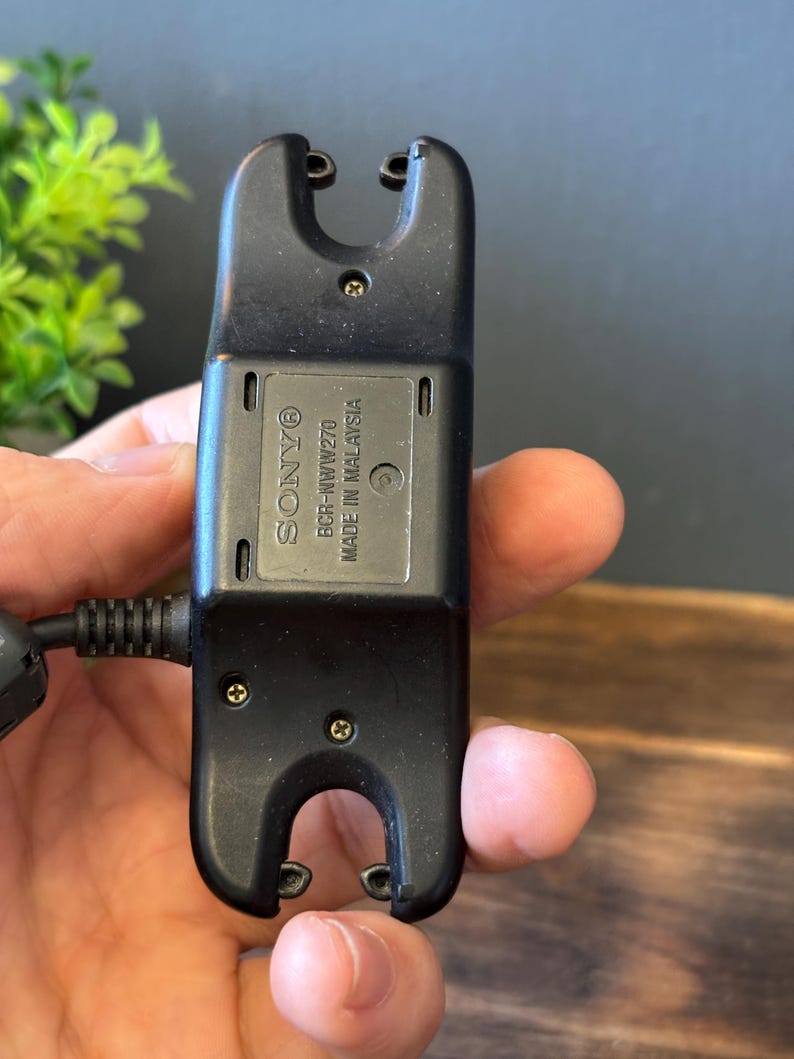May include: A black Sony BCR-NWW270 charging cradle for a wearable device. The rectangular plastic unit features two U-shaped cutouts at the top and bottom, exposed screws, and text reading SONY BCR-NWW270 MADE IN MALAYSIA on the center panel.