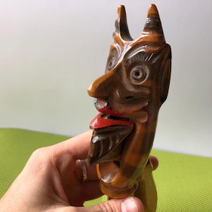Op de afbeelding: Een houten notenkraker gesneden in de vorm van een duivels gezicht. De notenkraker heeft een rode mond, een bruine baard en een bruin en zwart geschilderd gezicht.