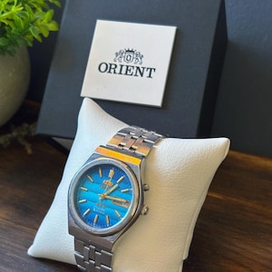 May include: An Orient wristwatch with a silver-tone metal link band and a vibrant blue gradient dial. The watch features gold-tone hour markers and hands, a day-date window at the three o'clock position, and the Orient logo printed on the dial.